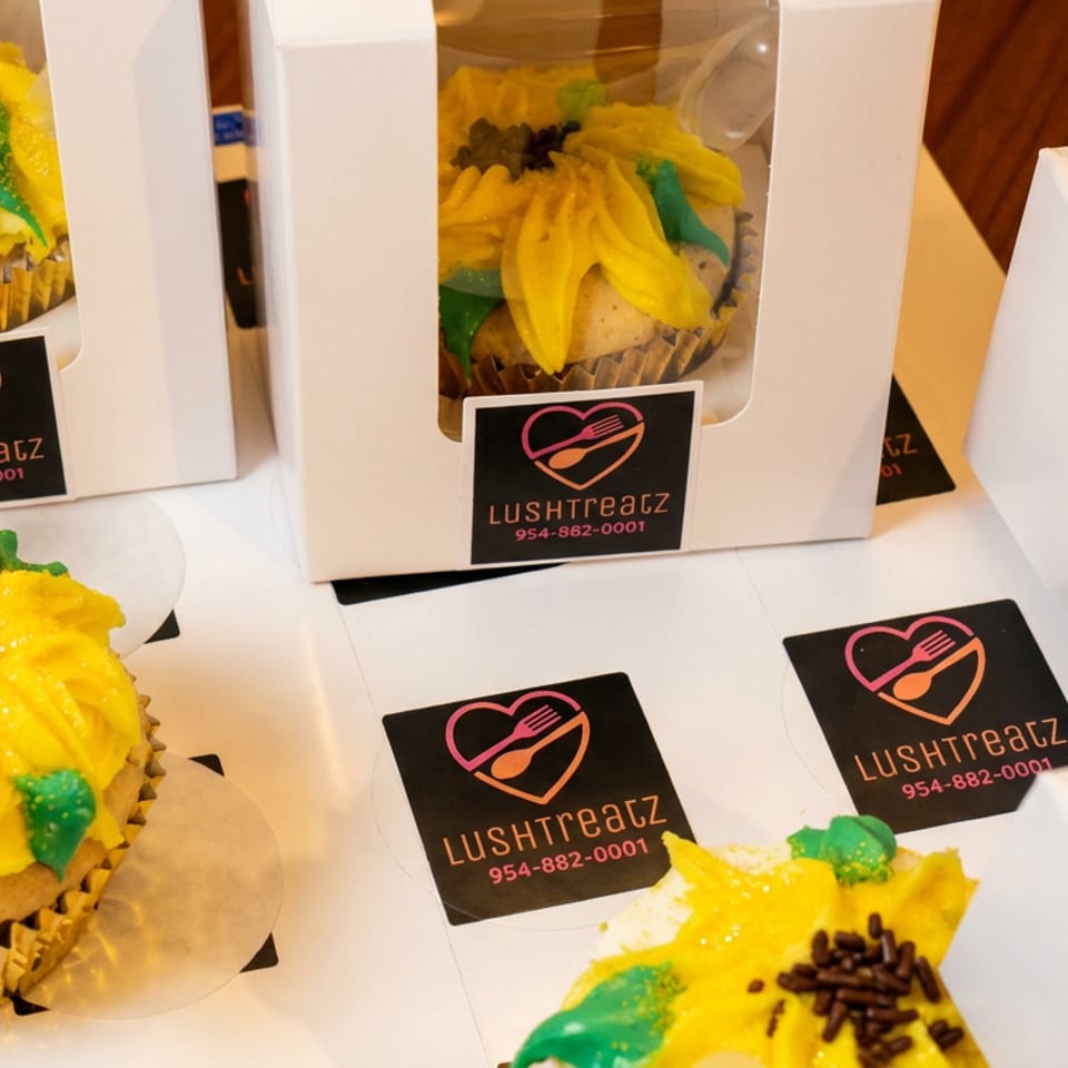 Several Lushtreatz cupcakes with yellow sunflower frosting and sprinkles, some packaged in white boxes.